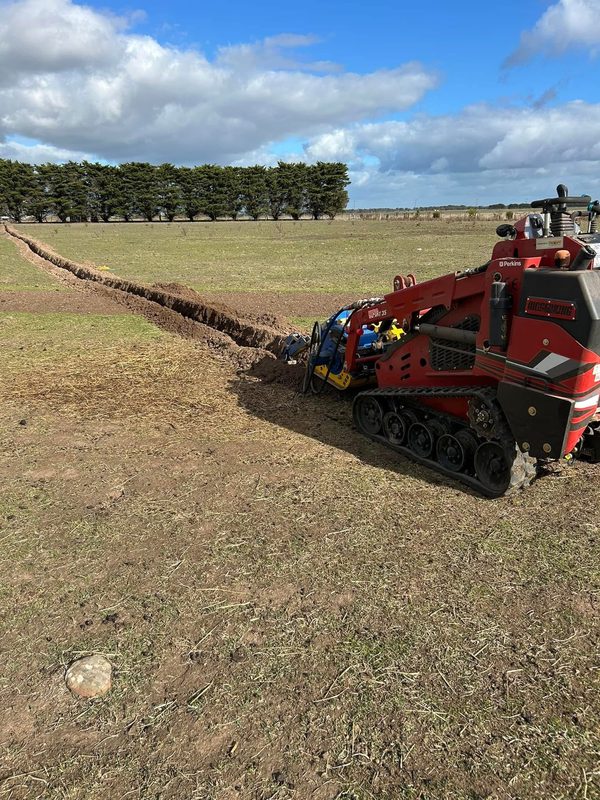 Red Darlington tracked mini loader with a trencher cutting a long, straight trench across a grassy paddock