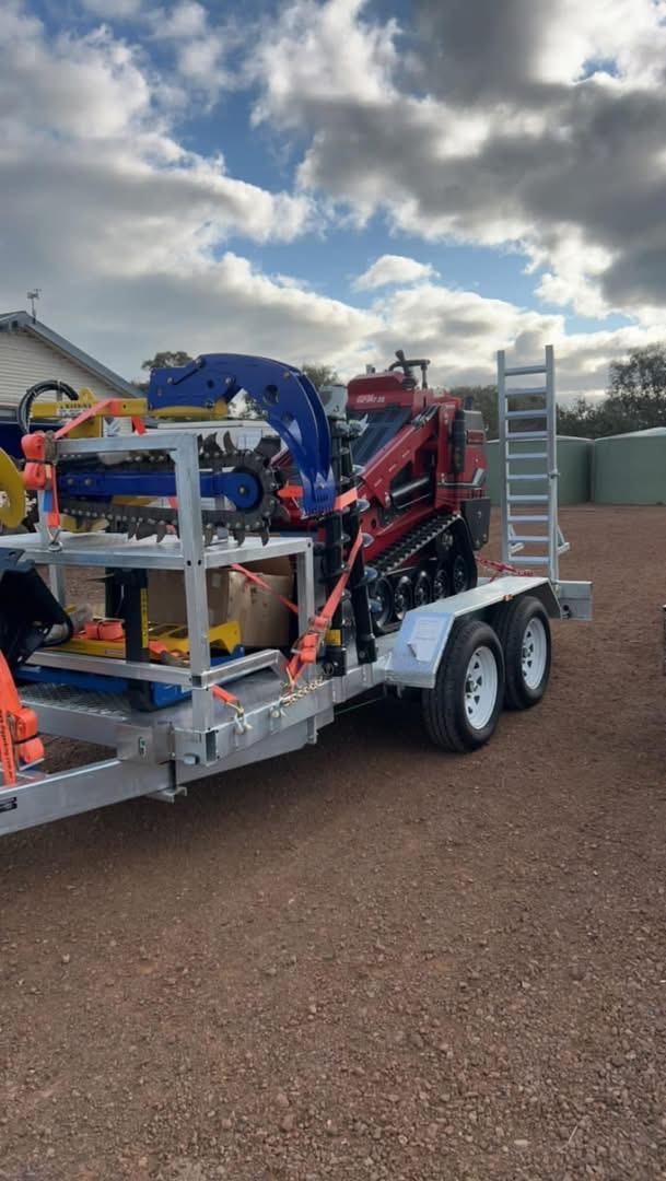 Red tracked mini loader loaded on a galvanised tandem plant trailer with the trencher and attachments on a rack