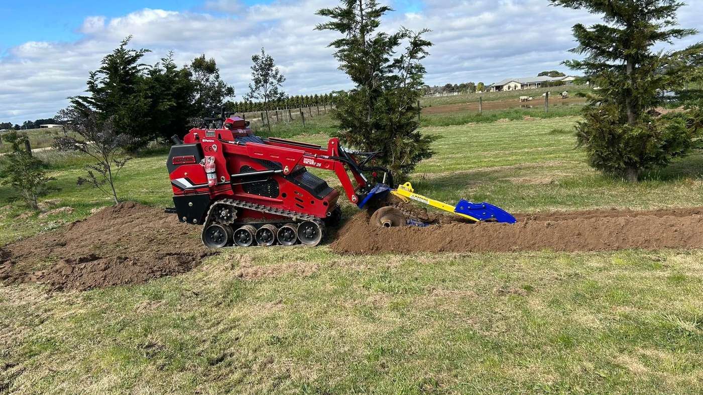 Red Darlington tracked mini loader with a trencher cutting a trench across a green lawn