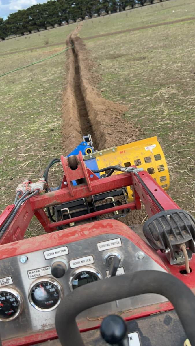 Operator's view from the mini loader controls as the yellow trencher cuts a straight trench across a paddock